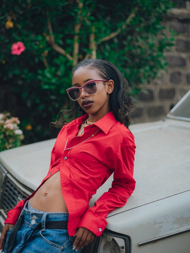 Young woman in red shirt and jeans leaning on car.